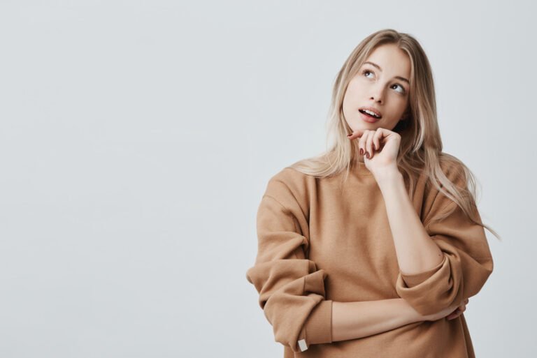 Portrait of pensive fair-haired dreamy hipster girl imagines something in her mind, looks upwards, being deep in thoughts, touching her chin. Attractive young blonde female dreams about vacation or weekends